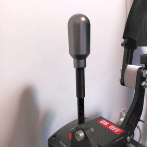 Puede incluir: Una perilla de palanca de cambios de metal gris con un eje negro. La palanca de cambios está unida a un simulador de carreras negro con un interruptor "ON OFF" rojo y un interruptor "KILL" negro.