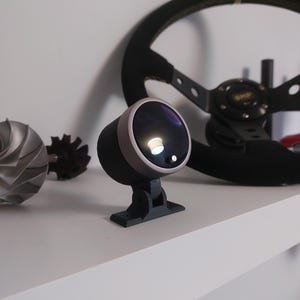 Puede incluir: Un medidor negro y gris con un bisel gris claro y una base negra. Un volante negro con costuras amarillas está en el fondo. También se ven una turbina plateada y un engranaje negro.