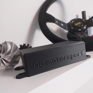 Puede incluir: Se muestra un componente rectangular negro con el texto "DE motorsport", junto con un volante negro con costuras amarillas y una turbina plateada. El volante tiene la marca "OMP". Los artículos están sobre una superficie blanca.