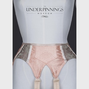 May include: A pink satin garter belt with white and gray lace trim. The garter belt has four straps and is decorated with a diamond-shaped pattern. The text "THE UNDERPINNINGS MUSEUM" is visible at the top of the image.