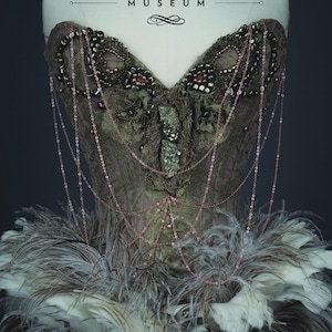 May include: A green and brown corset with beaded embellishments and chains, worn on a mannequin. The corset is adorned with feathers and the text "THE UNDERPINNINGS MUSEUM" and "GILDED LILIES" are visible.