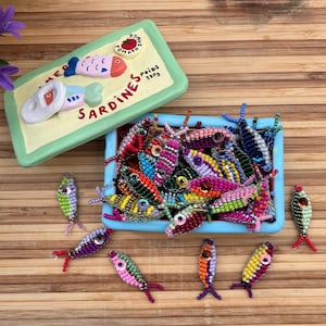 Könnte beinhalten: Eine blaue und weiße Schachtel mit den Worten "Sardines" und "Poids 130g" darauf. Die Schachtel ist geöffnet und mit bunten Perlenfischen gefüllt. Es liegen auch mehrere Perlenfische um die Schachtel herum.