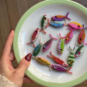 Könnte beinhalten: Eine Sammlung von bunten Perlfischen in Rosa, Grün, Gelb und Lila, angeordnet auf einem weißen Teller mit grünem Rand. Die Fische haben detaillierte Perlenarbeiten und kleine, dekorative Akzente.