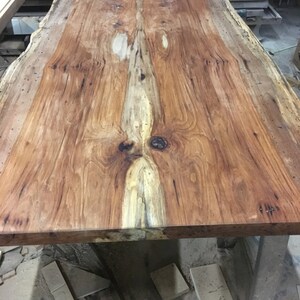 May include: A large, rustic wooden table top with a natural wood finish. The wood has a unique grain pattern and a light brown color. The table top is made of a single piece of wood and has a live edge.