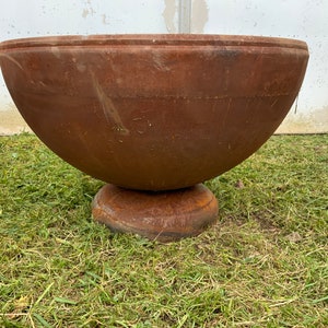 May include: A large, rusty metal bowl with a wide, rounded base. The bowl is sitting on a bed of green grass.