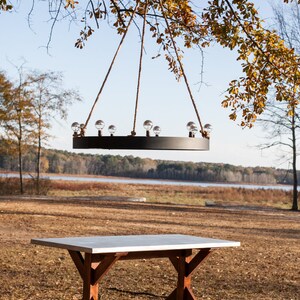 May include: A black circular chandelier with clear globe bulbs hangs from ropes attached to a tree branch. Below, a rectangular table with a light gray top and wooden trestle legs sits on the ground. The scene is outdoors with a lake in the background.
