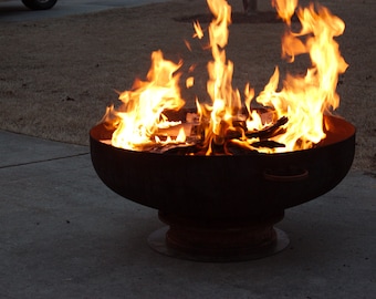 May include: A large metal fire pit with a wide, round bowl and a base. The fire pit is filled with burning wood and flames are rising high into the air.