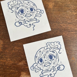 May include: Two blue ink prints of a moon face with large eyes and a mouth, surrounded by clouds. The moon face is stylized and has a textured surface.