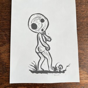 May include: Black and white line art print of a stylized human figure with a large, round head and two eyes. The figure is walking with its arms at its sides. The figure is standing on a patch of grass.
