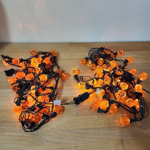 May include: Two strings of Halloween-themed orange pumpkin lights with black wiring. The pumpkins have carved faces and are lit, creating a warm glow. The lights are on a wooden surface.