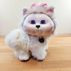 May include: A vintage white plush cat doll with a painted face, purple eyes, and a pink bow. The doll has a fluffy body and a black button on its collar. The cat's face has purple accents.