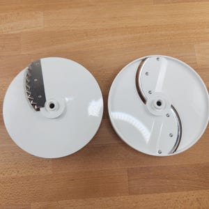 May include: Two white, circular food processor blades with stainless steel cutting edges. One blade has a serrated edge, and the other has a curved blade. Both have a central hub for attachment. The blades are on a wooden surface.