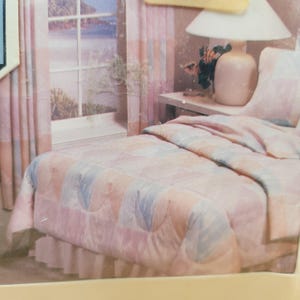 May include: A bed with a pastel-colored comforter, pillows, and a bed skirt. The comforter has a quilted pattern in shades of pink, blue, and peach. Matching curtains frame a window overlooking a seascape. A lamp sits on a bedside table.