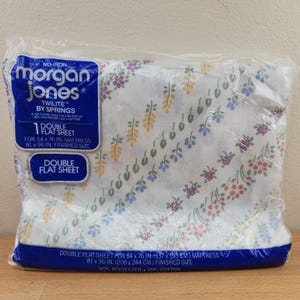 May include: A packaged double flat sheet by Morgan Jones. The sheet is white with a floral pattern in shades of purple, pink, yellow, and blue. The package is labeled for a 54 x 76 inch mattress, with a finished size of 81 x 96 inches.
