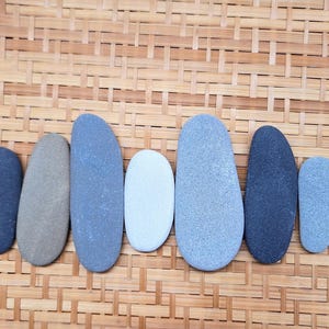 May include: Seven smooth, oval-shaped river stones arranged in a horizontal row. The stones vary in colour, including shades of dark charcoal, light grey, tan, and pale blue-grey, each featuring a matte, natural texture and rounded edges.