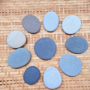 May include: A collection of ten smooth, oval-shaped stones arranged in a circular pattern. The stones vary in colour, featuring shades of light grey, medium grey, dark grey, and blue-grey. Each stone has a matte texture and rounded edges.