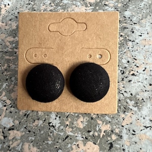 Puede incluir: Un par de pendientes de botón redondos cubiertos de tela negra. Los pendientes están sujetos a una tarjeta de exhibición de pendientes de cartón marrón. El fondo es una superficie moteada gris y blanca.