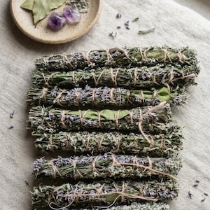 Könnte beinhalten: Sieben Bündel aus getrocknetem Lavendel und grünen Kräutern, die mit dünnem Garn zusammengebunden und in einer Reihe angeordnet sind. Eine kleine, runde, gesprenkelte beige Keramikschale enthält getrocknete Lorbeerblätter und zwei violette Amethyststeine.