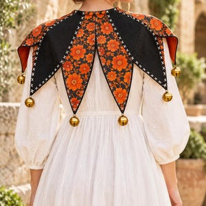 May include: A white long-sleeved dress paired with a black and orange floral print jester-style collar. The collar features pointed panels, white cross-stitch detailing along the edges, and gold-toned metal bells attached to each of the six points.