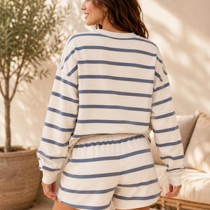 May include: A two-piece lounge set consisting of a long-sleeved crewneck sweatshirt and matching shorts. Both pieces feature a horizontal striped pattern in muted blue on an off-white background, with a relaxed fit and ribbed cuffs.