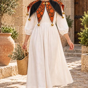 May include: A long-sleeved cream-colored dress paired with a black and orange floral-patterned collar featuring gold-toned jingle bells. The collar is secured with a gold clasp. A woven straw basket is held in one hand, and brown sandals are worn.