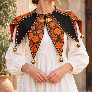 May include: A black and orange floral print capelet featuring a pointed collar design with white cross-stitch detailing along the edges. The garment is secured at the neck with a gold-toned metal clasp and adorned with small gold jingle bells.