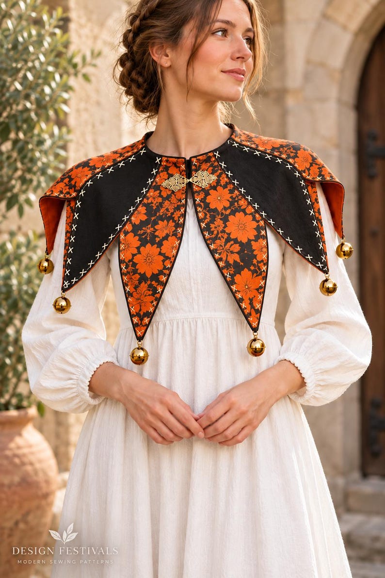 May include: A black and orange floral print capelet featuring a pointed collar design with white cross-stitch detailing along the edges. The garment is secured at the neck with a gold-toned metal clasp and adorned with small gold jingle bells.