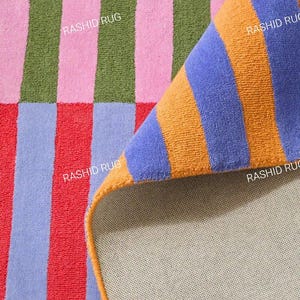 Può includere: Un tappeto rettangolare con un motivo a righe nei colori verde oliva, rosa, azzurro, rosso e arancione. Un angolo è ripiegato per rivelare il retro beige chiaro. Il testo RASHID RUG è stampato più volte sulla superficie del tappeto.