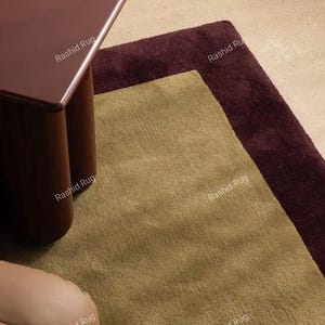 May include: A rectangular area rug featuring a large central panel in a muted olive green hue, bordered by a deep burgundy frame. The rug has a short, dense pile texture and is partially covered by a dark maroon table with rounded legs.