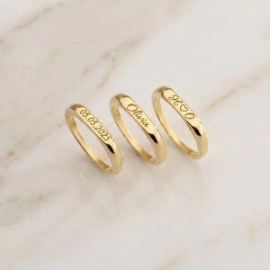 May include: Three gold-toned rings with smooth, rounded bands. Each ring features a custom engraving: the first displays the date 05.05.2025, the second shows the name Olivia, and the third displays the letters H and O separated by a heart symbol.