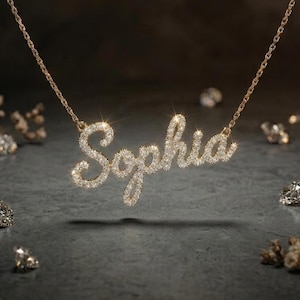 May include: A gold-toned necklace featuring a cursive nameplate pendant that spells out the name Sophia. The letters are encrusted with numerous small, clear, sparkling stones that catch the light, suspended from a delicate matching gold chain.