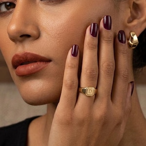 May include: A gold signet ring featuring a polished finish and a central square-shaped face engraved with the letter D. The ring is worn on the ring finger, showcasing a smooth band and a flat, geometric top surface with clean, defined edges.