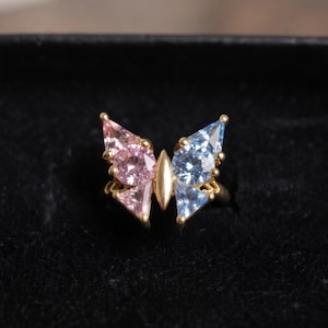 May include: A gold ring featuring a butterfly design with two pink faceted gemstones on the left wing and two light blue faceted gemstones on the right wing, centered by a smooth gold marquise-shaped piece in the middle.