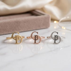 Könnte beinhalten: Drei Ringe in einer Reihe angeordnet, jeder mit einem stilisierten Monogramm der Buchstaben G und D. Der erste Ring ist goldfarben, der zweite roségoldfarben und der dritte silberfarben. Jeder Ring hat eine dünne Ringschiene und eine glatte Oberfläche.