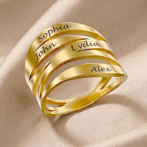 May include: A gold-colored ring with four bands, each engraved with a name: Sophia, John, Lydia, and Alex. The ring has a modern design with a polished finish, set against a neutral background.