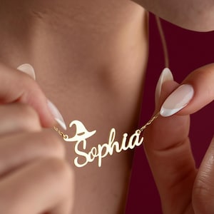May include: A gold-toned metal necklace featuring a cursive nameplate that reads Sophia. The nameplate is accented with a small, stylized witch hat icon positioned above the letter S. The pendant is attached to a delicate gold-toned chain.