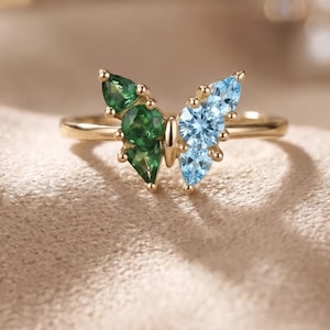 May include: A gold ring featuring a butterfly-shaped centerpiece. The left wing is composed of three green gemstones in pear and round cuts, while the right wing consists of three light blue gemstones in pear and round cuts, set in gold.