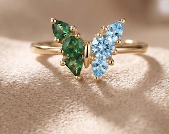 May include: A gold ring featuring a butterfly-shaped centerpiece. The left wing is set with three green gemstones in pear and round cuts, while the right wing features three light blue gemstones in pear and round cuts.