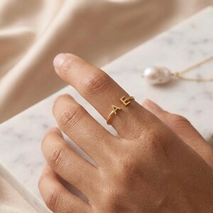 May include: A gold-toned ring featuring two capital letters, A and E, positioned at the open ends of the band. The ring has a polished metallic finish and a simple, thin circular design that rests on the finger, showcasing the two distinct letters.