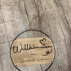 Könnte beinhalten: Rundes Holzschild mit den Worten "Willkommen bei Jonas & Sophie" in schwarzer Schrift. Das Schild wird von einem dünnen, braunen Seil gehalten und von einem schwarzen Metallring umrahmt. Dekorative Herzen sind enthalten.