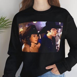 Op de afbeelding: Zwart sweatshirt met een rechthoekige print van een filmstill met twee mensen met cowboyhoeden. Het sweatshirt wordt gedragen door een persoon die een blauwe spijkerbroek draagt.