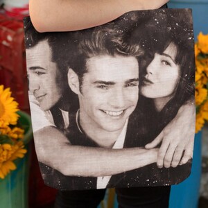 May include: A black and white tote bag featuring a vintage photograph of three people. The bag is held by a person in a light green top, surrounded by sunflowers and other colourful flowers in a flower shop.