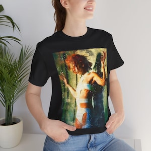 May include: Black t-shirt featuring a watercolor painting of a person with red hair and white clothing. The artwork has a green and yellow background with a moon and stars.