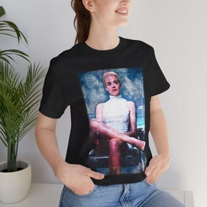 May include: Black t-shirt featuring a portrait of a woman in a white turtleneck and dress. The image has a blue and white background. The woman is seated with her legs crossed. The shirt is made of a soft material.