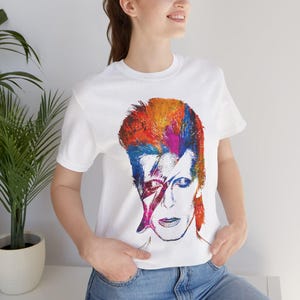 Könnte beinhalten: Weißes T-Shirt mit einem farbenfrohen Porträt eines Musikers mit einem Blitz auf dem Gesicht. Das Kunstwerk verwendet eine lebendige Palette aus Rot, Orange, Blau und Lila. Das Shirt ist ein klassischer Rundhalsausschnitt.