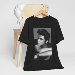 May include: Black t-shirt featuring a black and white portrait of a man with dark hair and a bracelet. The image is centered on the shirt, which is hanging on a wooden hanger. A white baseball cap rests on a wooden chair.