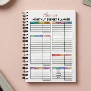 May include: A spiral-bound monthly budget planner with the text "Aleena's MONTHLY BUDGET PLANNER". The planner has sections for income, expenses, bills, savings, and debt. The planner is on a pink surface.
