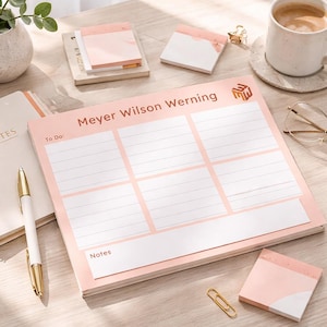 May include: A desk setup featuring a pink and white notepad with the text "Meyer Wilson Werning" and a "To Do" section. A spiral-bound notebook, pen, sticky notes, and a cup of coffee are also present. A small plant in a white pot adds a touch of greenery.