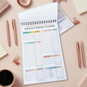 May include: A spiral-bound monthly budget planner with the text "Montgomery Family Monthly Budget Planner." The planner has sections for income, expenses, bills, debt, and savings. Rose gold pens and paper clips are scattered around the planner.