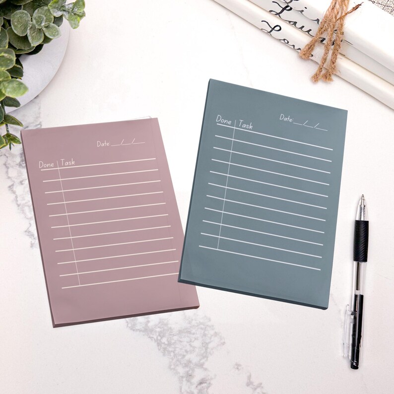 May include: Two rectangular notepads, one mauve and one teal, with white lines and the words "Done | Task" and "Date" printed on them. A black and silver pen rests on the white surface next to the teal notepad. The notepads are for organizing tasks.